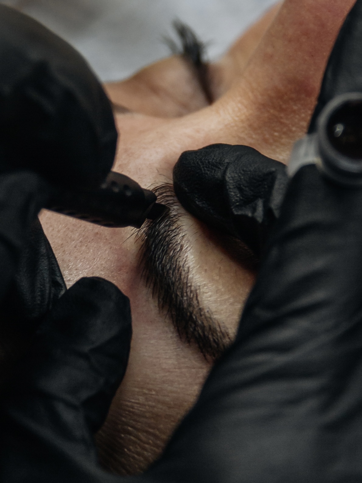 Close-up of gloved hands drawing a microblading blade along the upper edge of a brow, with a pigment cap resting alongside
