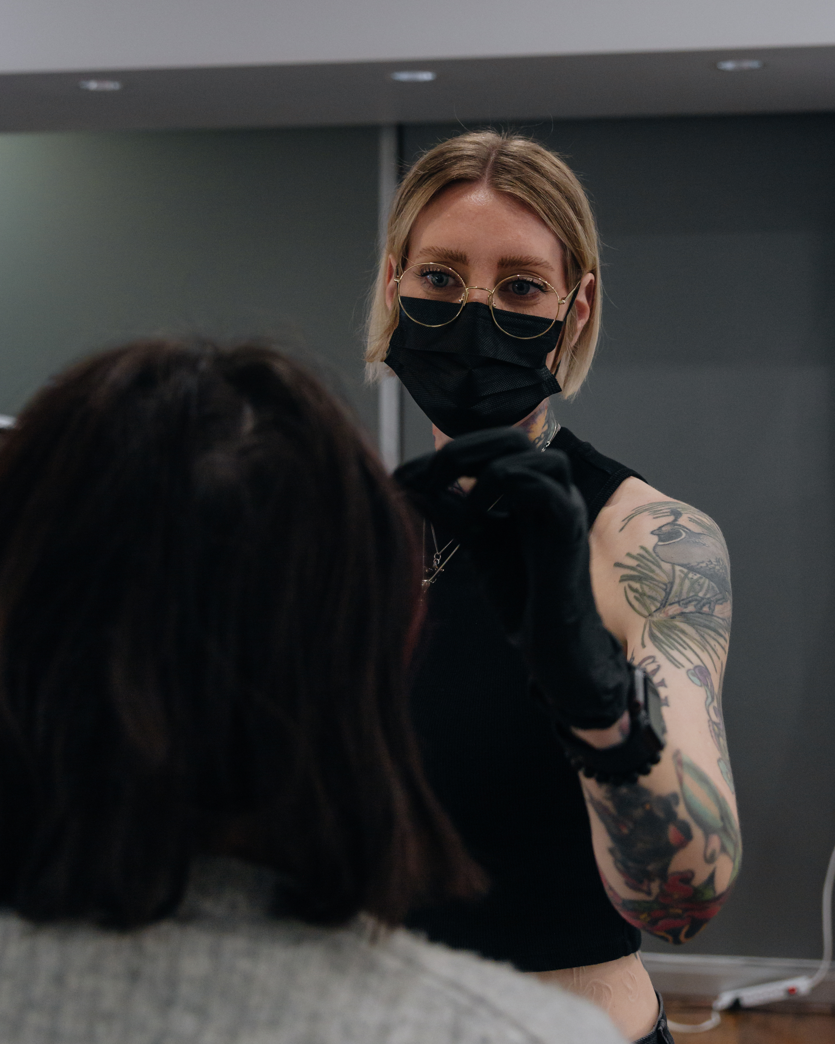 Sara B. looking toward the camera during an appointment, wearing a black mask, wire-rimmed glasses and black gloves, with a client seated in the foreground
